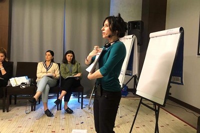 Adriana Leigh G. standing up presenting to a group of training participants (2 women sitting down are seen in the picture).