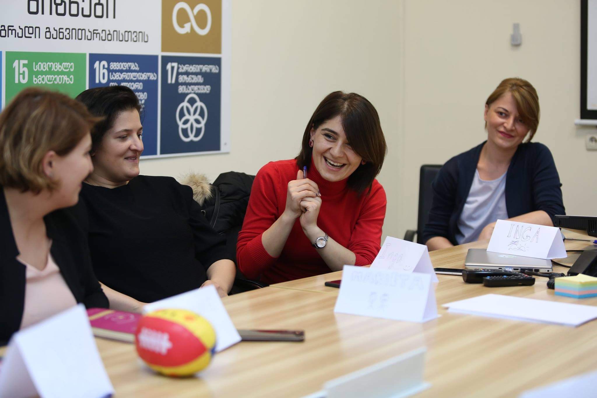 Photo of women at a table smiling with a photo of the sustainable development goals in Georgian language behind them