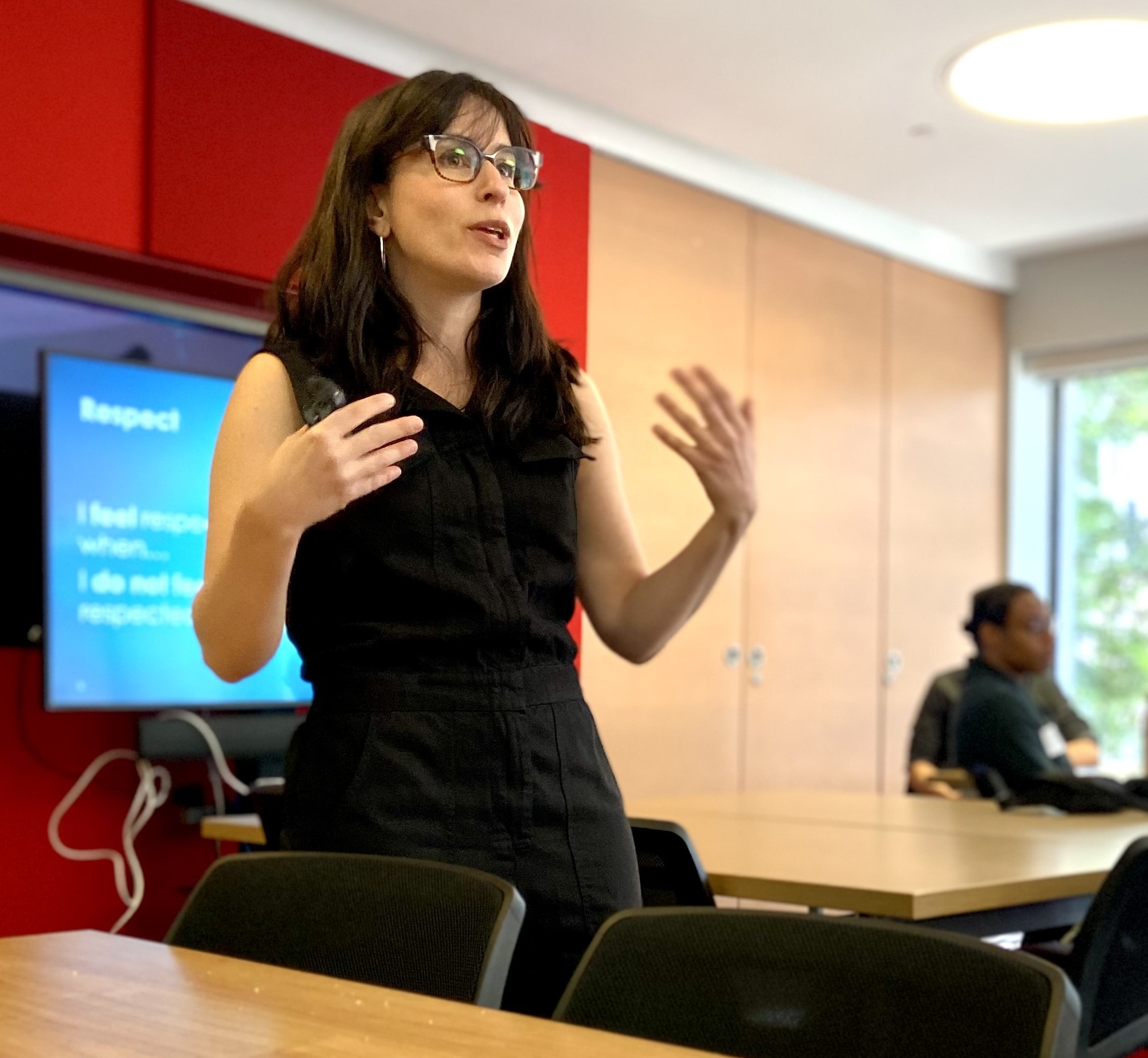 Woman in black jumpsuit with long brown hair and glasses standing up giving a presentation