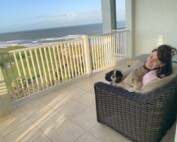 Picture of white Jewish woman with long brown hair smiling at the camera sitting on a chair on a balcony by the water. She is joined by her dog, a black and white floppy-eared little dog.