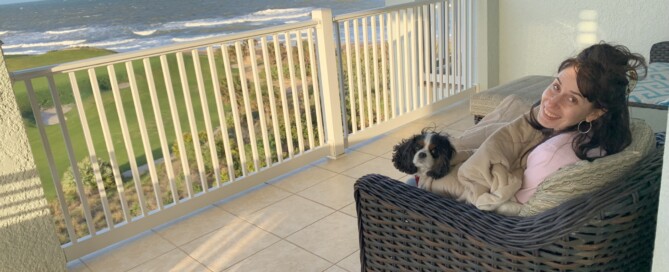 Picture of white Jewish woman with long brown hair smiling at the camera sitting on a chair on a balcony by the water. She is joined by her dog, a black and white floppy-eared little dog.
