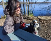 Photo of a white Jewish woman smiling at her dog. She is sitting on a bench by the water and wearing sunglasses.