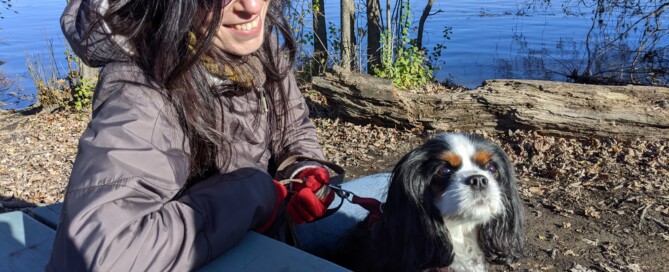 Photo of a white Jewish woman smiling at her dog. She is sitting on a bench by the water and wearing sunglasses.