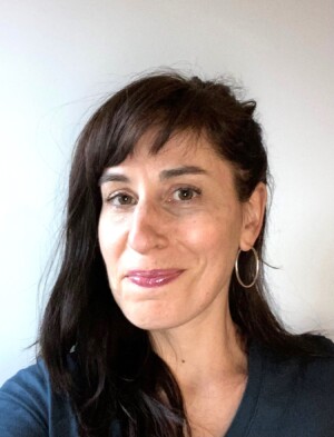 Picture of a white Jewish woman with long brown hair and bangs smiling at the camera. She is wearing a dark blue t-shirt.