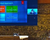 Photo of a white Jewish woman with glasses and dark brown hair on a screen giving a virtual presentation on zoom - there is a powerpoint with the words "safety and security" in an orange box , "confidentiality" in a green box, "respect" in a light blue box and "non-discrimination" in a dark blue box.