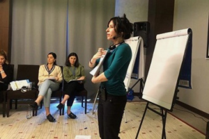 Photo of a white Jewish woman with brown hair giving a presentation smiling. She is wearing black pants and a teal top.