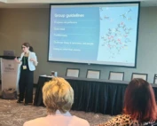 Picture of a Jewish white woman giving a presentation. She has long brown hair in a ponytail wearing glasses and a turquoise blazer and black pants. She has her hands up in an active gesture and is smiling.