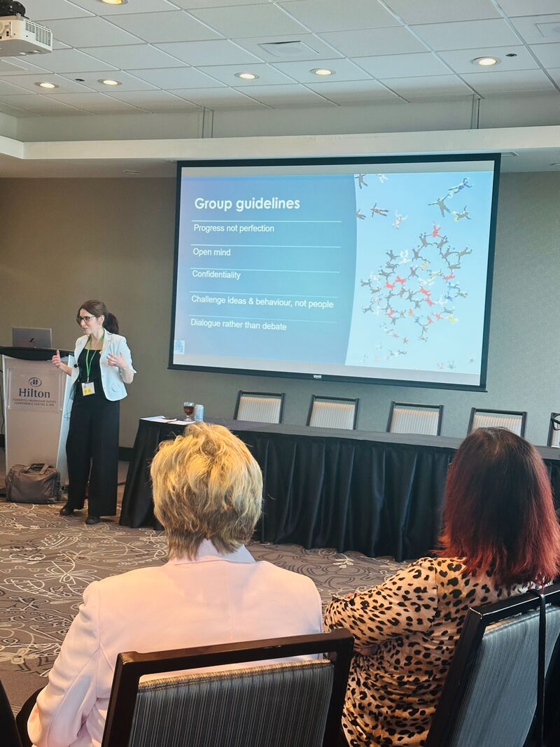 Picture of a Jewish white woman giving a presentation. She has long brown hair in a ponytail wearing glasses and a turquoise blazer and black pants. She has her hands up in an active gesture and is smiling.