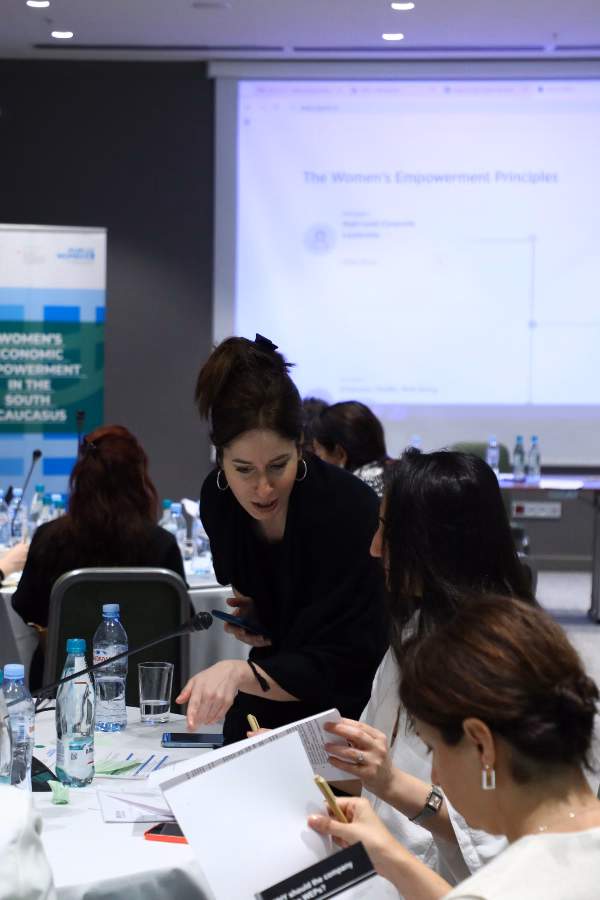 A woman dressed in black in a conference room, giving feedback to training attendees seated around a table.