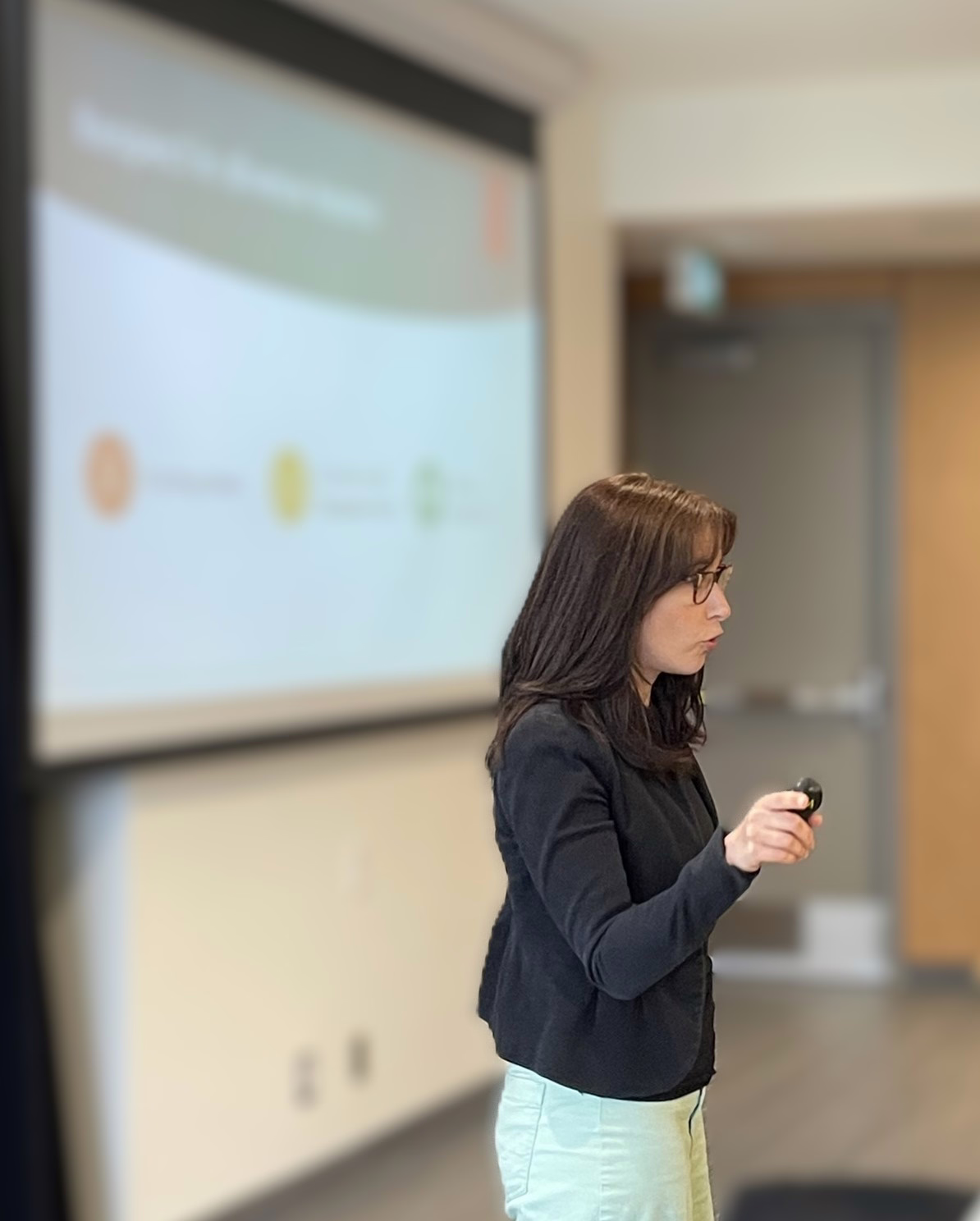 EN: A woman presenting at a conference, holding a clicker with a projected slide visible in the background. FR: Une femme donnant une présentation lors d'une conférence, tenant une télécommande avec une diapositive projetée visible en arrière-plan.