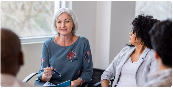 ENG: Photo of a woman with grey hair talking with others. FR : Photo d'une femme aux cheveux gris en train de discuter avec d'autres personnes.